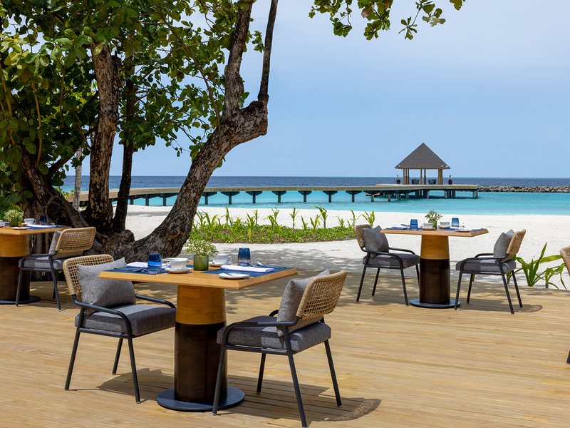 Restaurant tables under the trees, the beach and jetty beyond