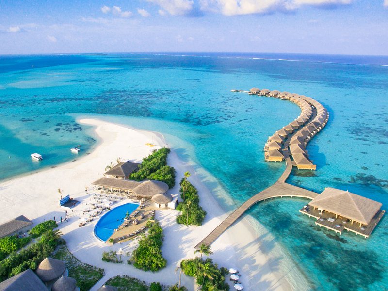 The island, its sandbank and a curve of overwater villas, from the air