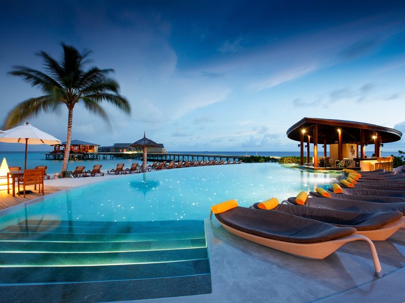 An infinity pool and loungers at dusk, with overwater villas beyond